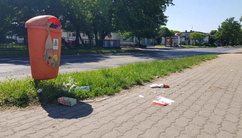 Śmieci, wysypujące się z pomarańczowego pojemnika na odpady w pasie drogowym Torunia